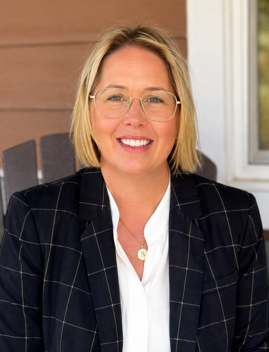 Smiling woman with short blond hair and large gold-rimmed glasses, wearing a navy plaid blazer on a porch.